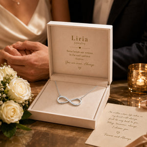 Silver infinity necklace in an open Liria jewelry box with a romantic message on a wooden table.