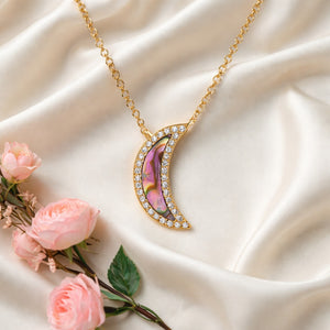 Gold necklace with a crescent moon pendant on a beige fabric background with pink roses.