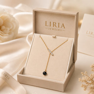 A gold Y-shaped necklace with a small clear stone and a single black bead pendant, presented in an open beige LIRIA JEWELRY box on a soft cream fabric background.