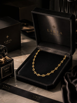A gold chain necklace displayed inside an open black LIRIA JEWELRY box, with matching branded packaging and a perfume bottle on a dark marble surface.