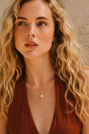 A close-up lifestyle shot of a woman with long, wavy blonde hair wearing a delicate gold necklace with a square white stone pendant, paired with a rust-brown top against a textured beige wall.