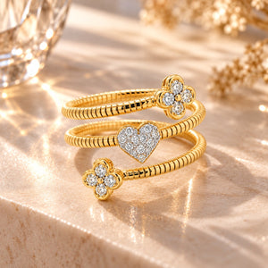 Three gold rings stacked together on a beige stone surface, featuring two flower-shaped settings and one heart-shaped pavé center, with sparkling clear stones and warm sunlight shadows.
