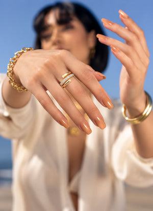 A close-up lifestyle shot of a model wearing gold rings and bracelets, with her hands raised in front of a soft-focus blue sky background.