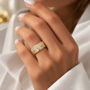 A close-up of a hand wearing a gold ring set with small clear stones, styled with a white satin sleeve in a softly lit setting.