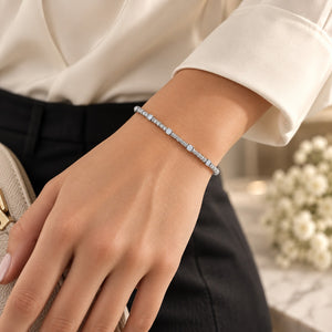 A delicate silver tennis bracelet with clear rectangular gemstones worn on a woman's wrist, paired with a cream blouse and black trousers.