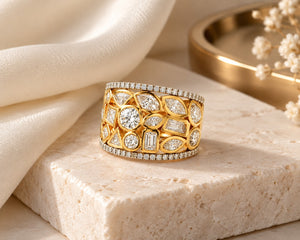 A wide yellow gold ring with multiple round and marquise-cut clear stones, displayed on a beige stone slab with draped cream fabric and small white flowers in the background.
