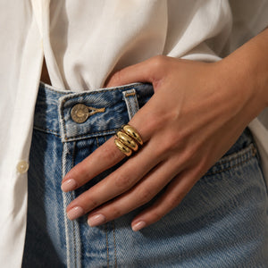 A close-up of a person's hand wearing a chunky gold ring with three coiled bands, styled with a white button-down shirt and blue denim jeans.