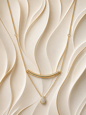 A gold layered necklace set displayed on a beige wavy textured background. The set features a gold bar necklace with a curved bar pendant, a delicate gold chain with a small teardrop-shaped crystal cluster pendant, and a thin gold chain with a small gold ring pendant.