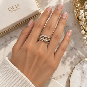 A close-up of a hand wearing three delicate rings from Liria Jewelry, with the brand's cream-colored box and a marble surface in the background.