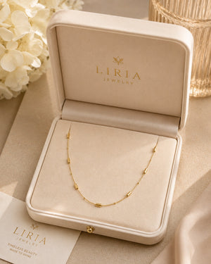 A delicate gold necklace with small elongated beads is displayed inside an open cream-colored LIRIA Jewelry presentation box, with floral and glass props in the background.