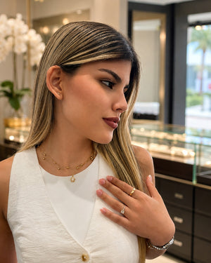 A woman in a white sleeveless top wearing delicate gold jewelry, including a layered necklace with a small gold heart pendant, a gold ring, and a silver ring with a clear gemstone, posing in a jewelry store.