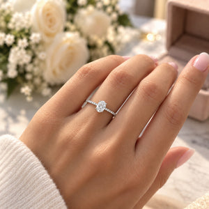 A close-up of a hand wearing a delicate silver ring with a round clear gemstone, set against a soft-focus background of white roses and a pink jewelry box.
