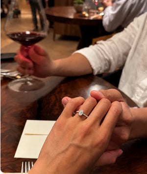 A close-up lifestyle shot of a person wearing a silver-toned diamond engagement ring on their ring finger, holding hands with another person in a warmly lit restaurant setting.