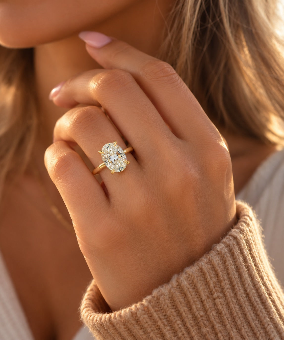 A close-up lifestyle shot of a woman's hand wearing a yellow gold ring with an oval-cut clear gemstone. The warm sunlight highlights her hand and sweater, and the ring is shown in a romantic, golden-hour setting.