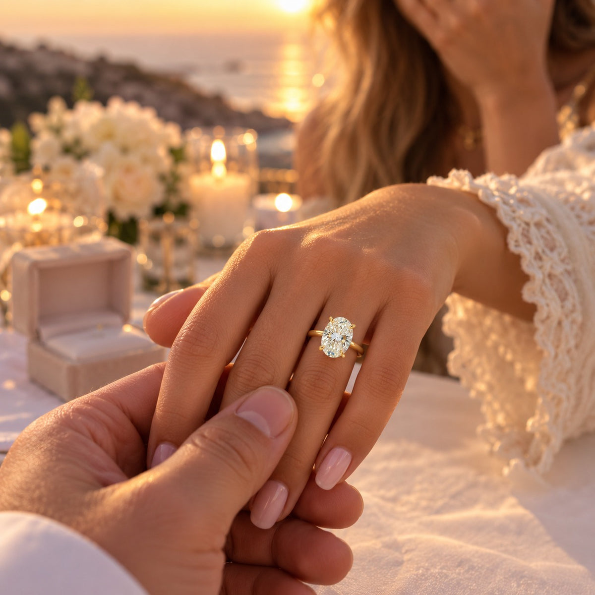 A close-up lifestyle shot of a woman's hand wearing a gold ring with a round brilliant-cut diamond, held by a man's hand in a romantic outdoor setting at sunset with candles and flowers.