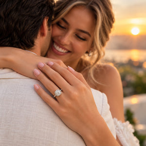 A woman smiling while hugging a man outdoors at sunset, wearing a silver engagement ring with a round diamond on her finger. The background is softly blurred with warm golden light.