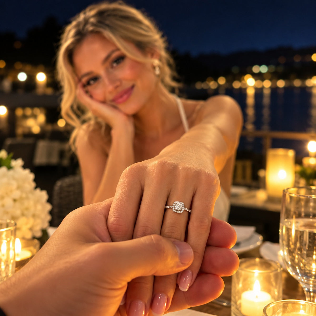 A woman at a candlelit dinner table extends her hand toward the camera, wearing a silver diamond ring with a round floral cluster setting. The background shows a softly blurred night-time waterfront setting with warm lights and water.