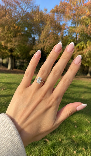 A hand with a French tip manicure wearing a silver ring with a rectangular clear gemstone, set against a blurred background of a park with autumn trees.