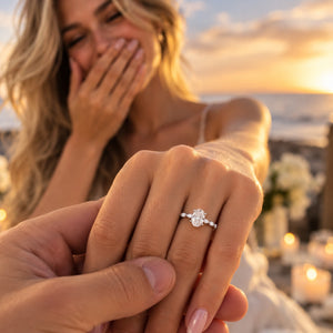 A close-up of a woman's hand wearing a silver-toned ring with a large round clear gemstone, while another person holds her hand. The background shows a softly blurred beach or outdoor setting at sunset with warm light and candles.