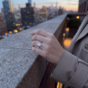 A close-up of a hand wearing a silver-toned ring with a large clear round gemstone, resting on a concrete ledge with a blurred city skyline at sunset in the background.