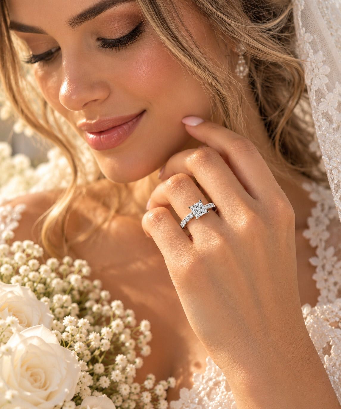 A close-up lifestyle shot of a bride wearing a diamond engagement ring on her finger, holding a bouquet of white roses. She is wearing a white lace wedding dress and veil.