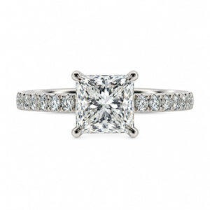 An engagement ring featuring a brilliant round-cut diamond center stone in a four-prong setting, with a pavé band set with small round diamonds, shown on a solid white background.