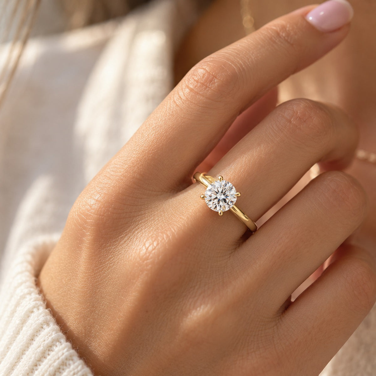 A close-up of a hand wearing a yellow gold solitaire engagement ring with a round brilliant-cut diamond. The hand has light pink manicured nails and is wearing a cream-colored knit sweater.