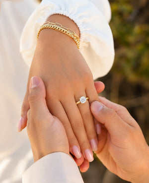 A close-up lifestyle shot of a gold engagement ring with a round-cut clear gemstone being worn on a hand, while another person holds it in place. The person wearing the ring has a gold chain bracelet on their wrist and is wearing a white sleeveless top.