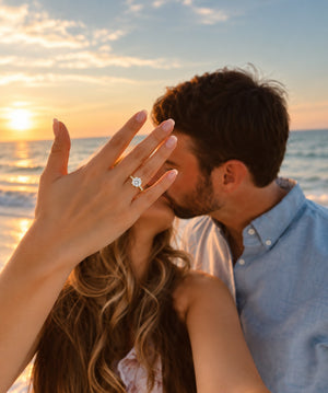 A woman on a beach at sunset shows an engagement ring on her finger as a man kisses her. The ring features a round brilliant-cut diamond set in a four-prong setting.