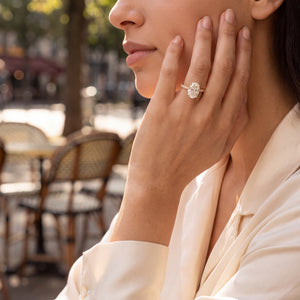 A close-up lifestyle shot of a woman wearing a diamond engagement ring with a halo setting on her hand, in an outdoor cafe setting.