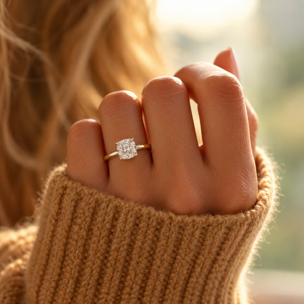 A close-up lifestyle shot of a hand wearing a gold engagement ring with a square-cut clear gemstone, set against a warm, softly blurred background with a person's blonde hair and a beige knit sweater sleeve visible.