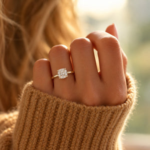 A close-up lifestyle shot of a hand wearing a gold engagement ring with a square-cut clear gemstone, set against a warm, softly blurred background with a person's blonde hair and a beige knit sweater sleeve visible.