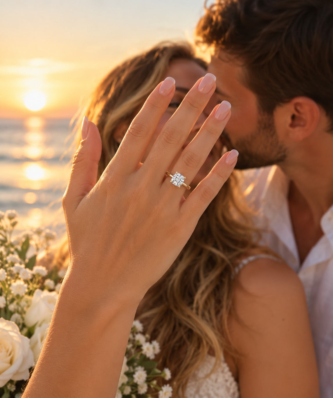 A close-up lifestyle shot of a woman's hand raised in front of her face, wearing a diamond engagement ring with a gold band. A man is kissing her cheek in the background, with a warm sunset over the ocean and white flowers visible.