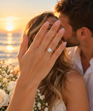 A close-up lifestyle shot of a woman's hand raised in front of her face, wearing a diamond engagement ring with a gold band. A man is kissing her cheek in the background, with a warm sunset over the ocean and white flowers visible.