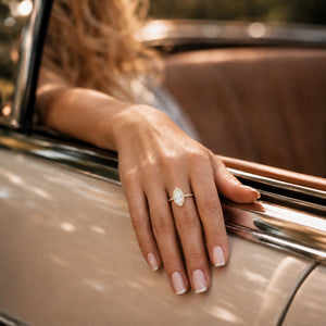 A close-up of a woman's hand resting on the door frame of a vintage car, wearing a delicate gold ring with a marquise-cut clear gemstone.