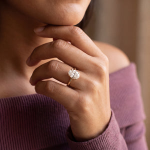 A close-up lifestyle shot of a woman wearing a yellow gold engagement ring with a round brilliant-cut diamond. The ring is shown on her ring finger, with her hand resting near her chin, wearing a mauve off-the-shoulder top.