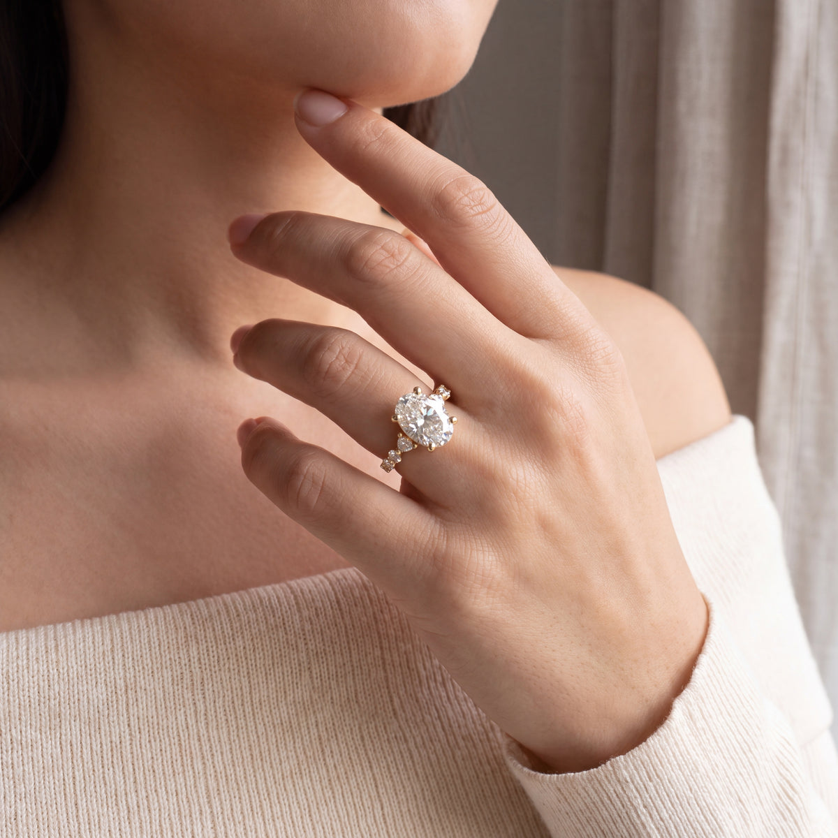 A close-up lifestyle shot of a woman wearing a delicate gold ring with a large round clear gemstone and small accent stones, set against a soft beige background.