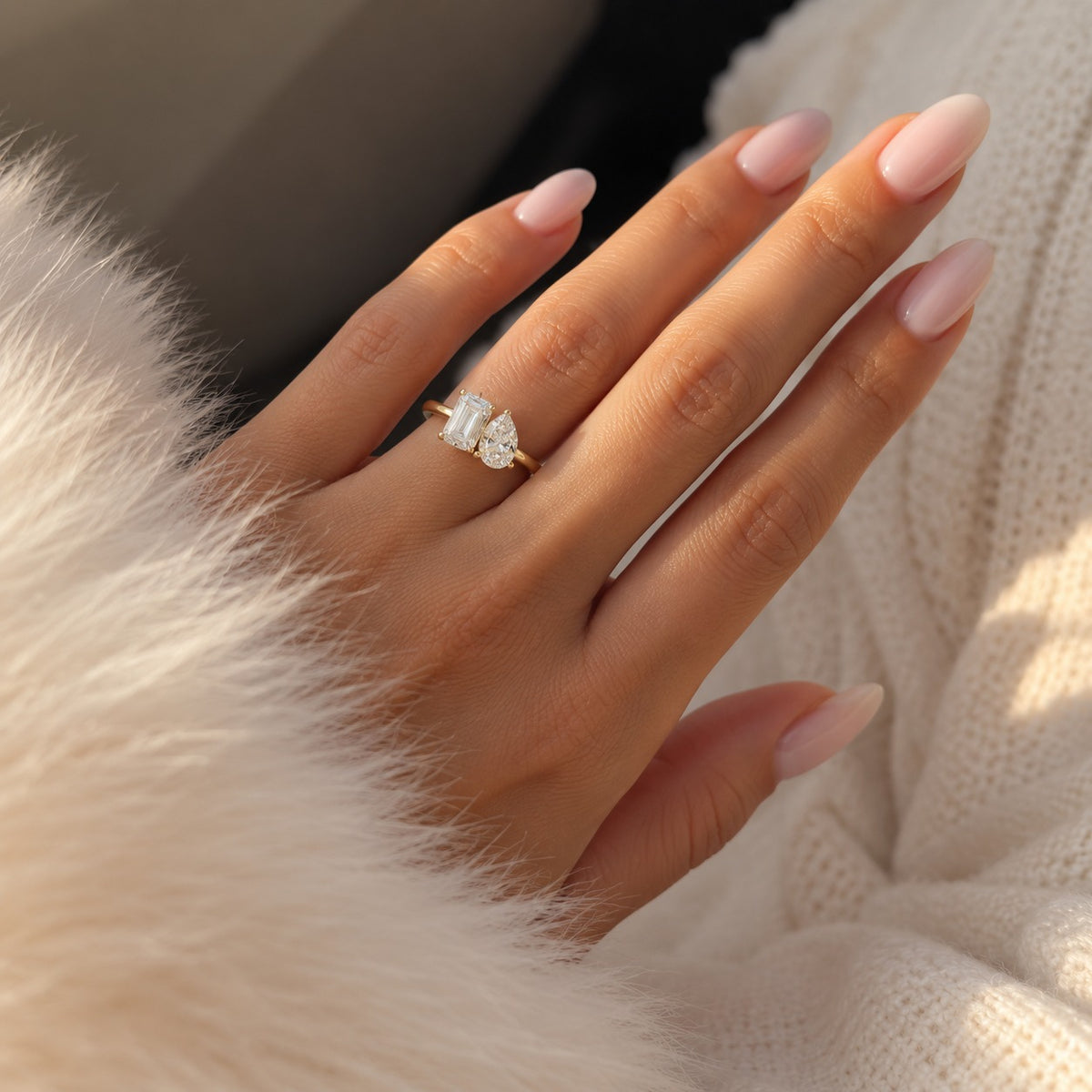 A close-up of a hand wearing a gold ring with a heart-shaped clear gemstone, styled with a cream knit sweater and fluffy cream faux fur.