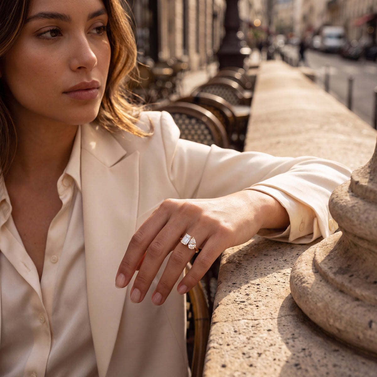A woman in a cream satin blazer and shirt leans on a stone ledge in an outdoor urban setting, wearing a silver-toned ring with a large clear gemstone.