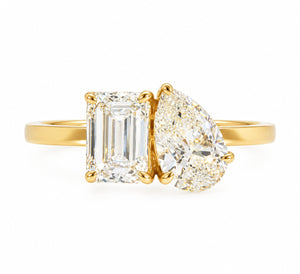 A yellow gold ring featuring an emerald-cut diamond on the left and a pear-cut diamond on the right, set against a plain white background.