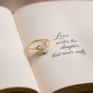 A gold ring with clear gemstones arranged in a floral shape, resting on the pages of an open book with elegant script text that reads 'Love writes the chapter that never ends.'.