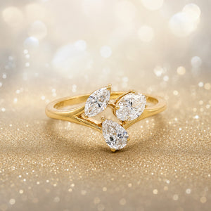 A gold ring featuring three marquise-cut clear gemstones arranged in a leaf-like cluster, photographed on a sparkling gold surface with a soft bokeh background.