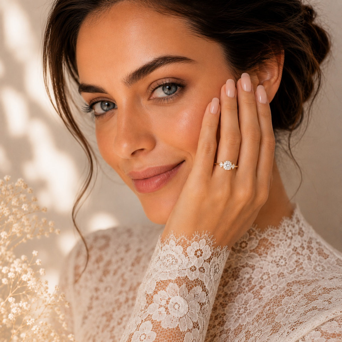 A close-up lifestyle shot of a woman with dark hair in an updo, wearing a delicate silver ring with a round clear gemstone on her ring finger. She is wearing a cream lace long-sleeve top and touching her cheek with her hand, set against a warm beige background with soft shadows.