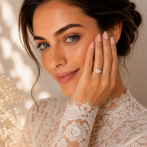 A close-up lifestyle shot of a woman with dark hair in an updo, wearing a delicate silver ring with a round clear gemstone on her ring finger. She is wearing a cream lace long-sleeve top and touching her cheek with her hand, set against a warm beige background with soft shadows.