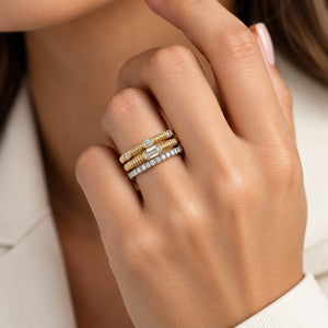 A close-up of a person's hand wearing three stacked rings: two textured yellow gold bands and one silver band with a central oval-cut diamond.