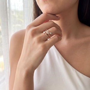 A close-up lifestyle shot of a woman wearing a delicate stack of two rose gold rings with small clear gemstones on her finger, set against a soft, bright background with sheer curtains.