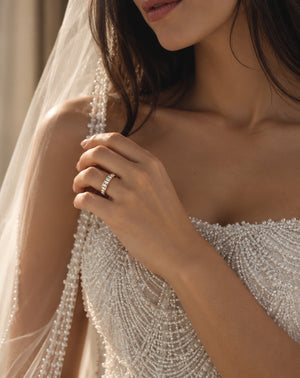 A close-up lifestyle shot of a bride wearing a delicate gold band ring set with small sparkling stones, paired with a white beaded wedding dress and veil.