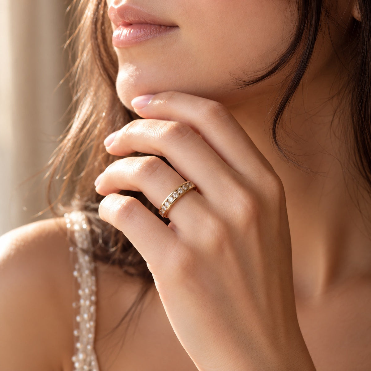 A close-up lifestyle shot of a woman wearing a delicate gold band ring set with small clear stones on her finger, with warm sunlight and a soft beige background.