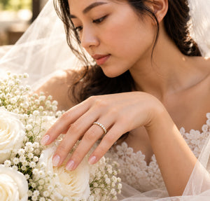 A close-up lifestyle shot of a bride wearing a delicate diamond band ring, gently touching a bouquet of white roses and baby's breath.
