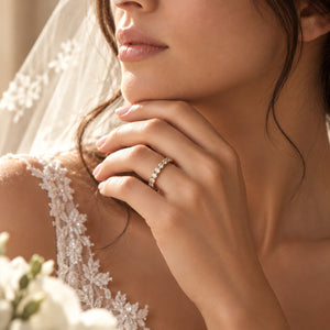 A close-up lifestyle shot of a bride wearing a delicate gold band ring set with small sparkling stones on her ring finger, wearing a white lace wedding dress and veil.